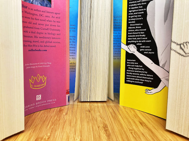 Three books sit on a light wood desk with their dust jackets and pages visible (all of the pages are still held together, almost as if the books were not even open. At the far left is a set of pages, followed by a right-hand flap in bright magenta. Next is a glimpse of a left-hand flap in sky blue with a streak of yellow, followed by pages, and then a glimpse of the right-hand flap with the same colors. Next is a left-hand flap with a bright yellow background, text enclosed in black, and two girls grabbing each other's hands, followed by more pages (these pages have a pair of holding hands printed on the edge).