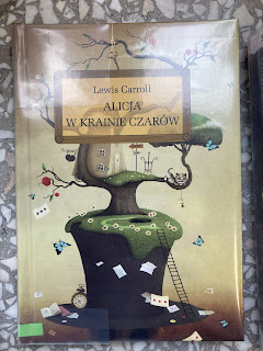 Na zdjęciu okładka książki na której widnieje tytuł ,,Alicja w krainie czarów" oraz autor Lewsi Carroll, okładka przedstawia drzewo na którym znajduje się domek, tło okładki jest beżowe.