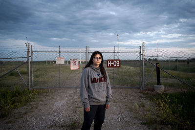 color photograph of young Native American woman Ella Weber in sweatshirt standing in front of gate to a nuclear silo on Fort Berthold Reservation