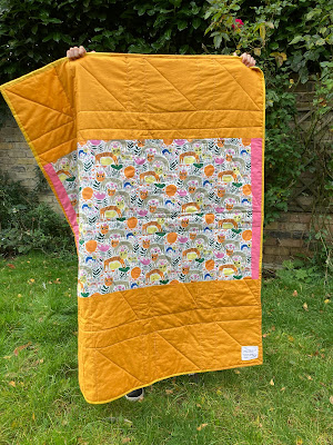 A golder yellow and tiger print quilt is being held up by a child.  They are standing on grass, with a brick wall behind.  You can only see the child's feet poking out under the quilt.