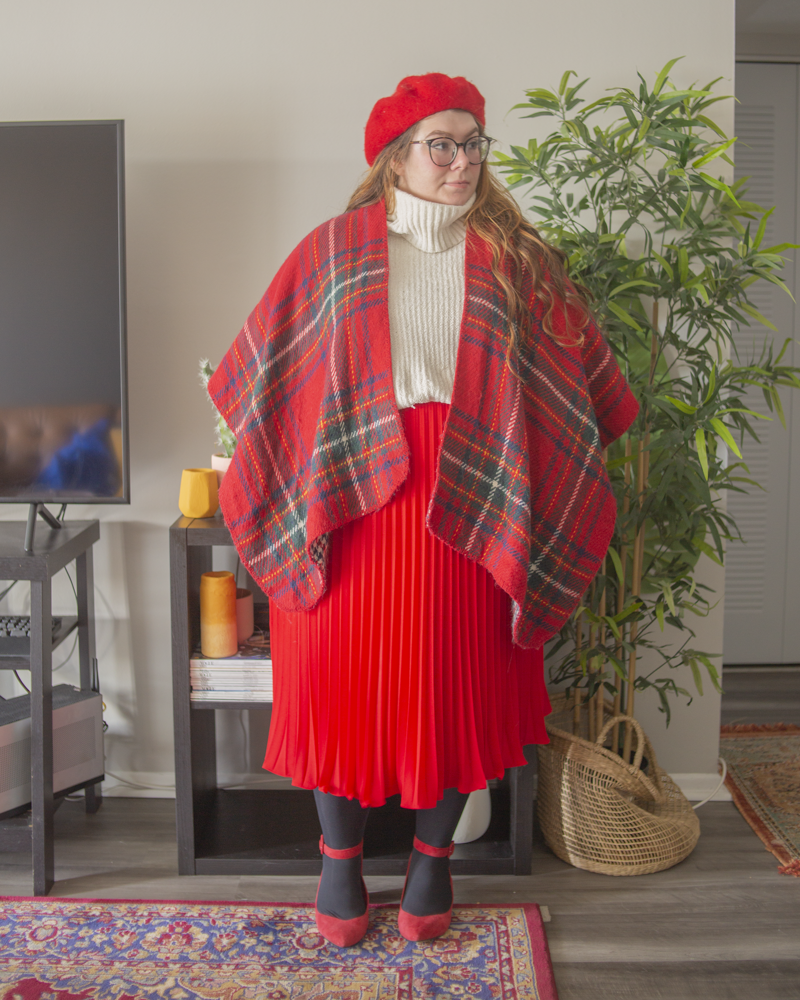 An outfit consisting of a red, white, green and blue plaid poncho over a white chenille turtleneck sweater, half tucked into a red pleated midi skirt and red ankle strap heels.