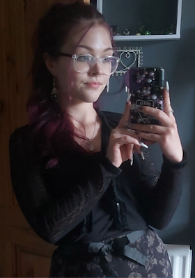 A selfie of Fern taken in a mirror. They have long dark purple hair tied up in a ponytail, glasses with transparent frames and are mostly dressed in black. Their phone case is bejewelled and they have long manicured nails painted with a white and black pattern.