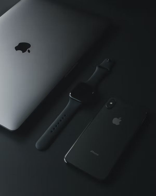 A black iPhone X showing the Apple logo and camera array. An Apple Watch screen up and with a closed silver MacBook next to it.
