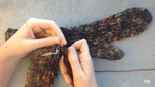 a textured crochet mitten laying flat; worked in dark yarn with flecks of yellow, orange, and red.  Someone's hands are visible, crocheting the matching mitten-in-progress.