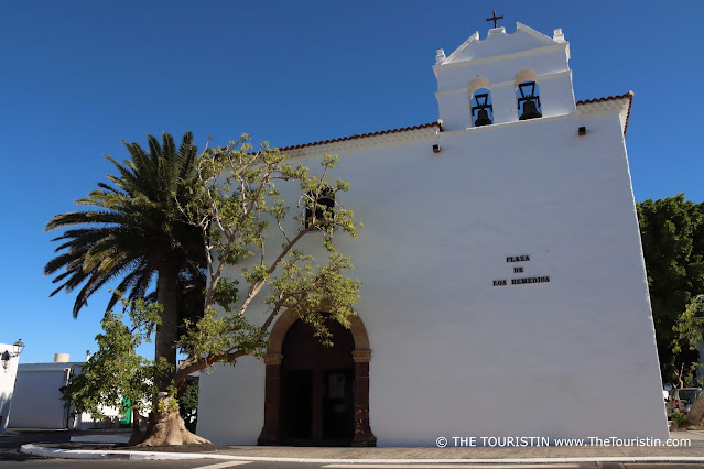 A large white cubic shaped church with a brown wooden entrance door and a large palm tree under a bright blue sky.
