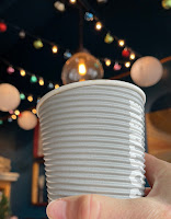a light grey compostable coffee cup being held up to the ceiling strung with Christmas lights