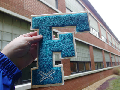 A hand holds up a felt letter "F" in front of a background of a two-story brick building with rows of windows.