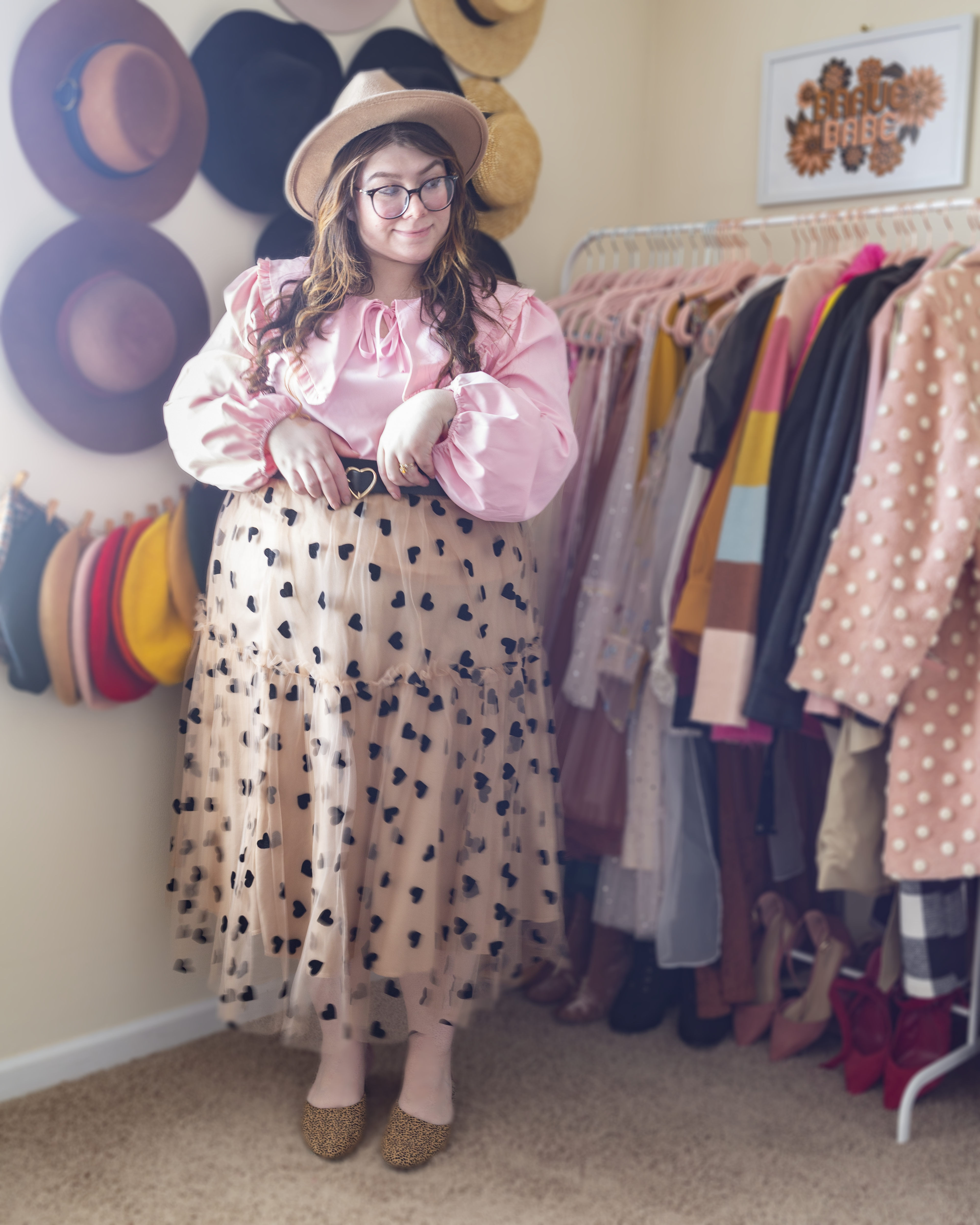 An outfit consisting of a pastel pink long bishop sleeve blouse with an oversized peter pan collar with ruffles on the hem, tucked into a sheer beige midi skirt with black velvet hearts and beige and black animal print heeled clogs.