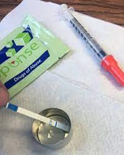 A fentanyl test strip in a a cap of water, with a needle and wrapper on a clean white towel.