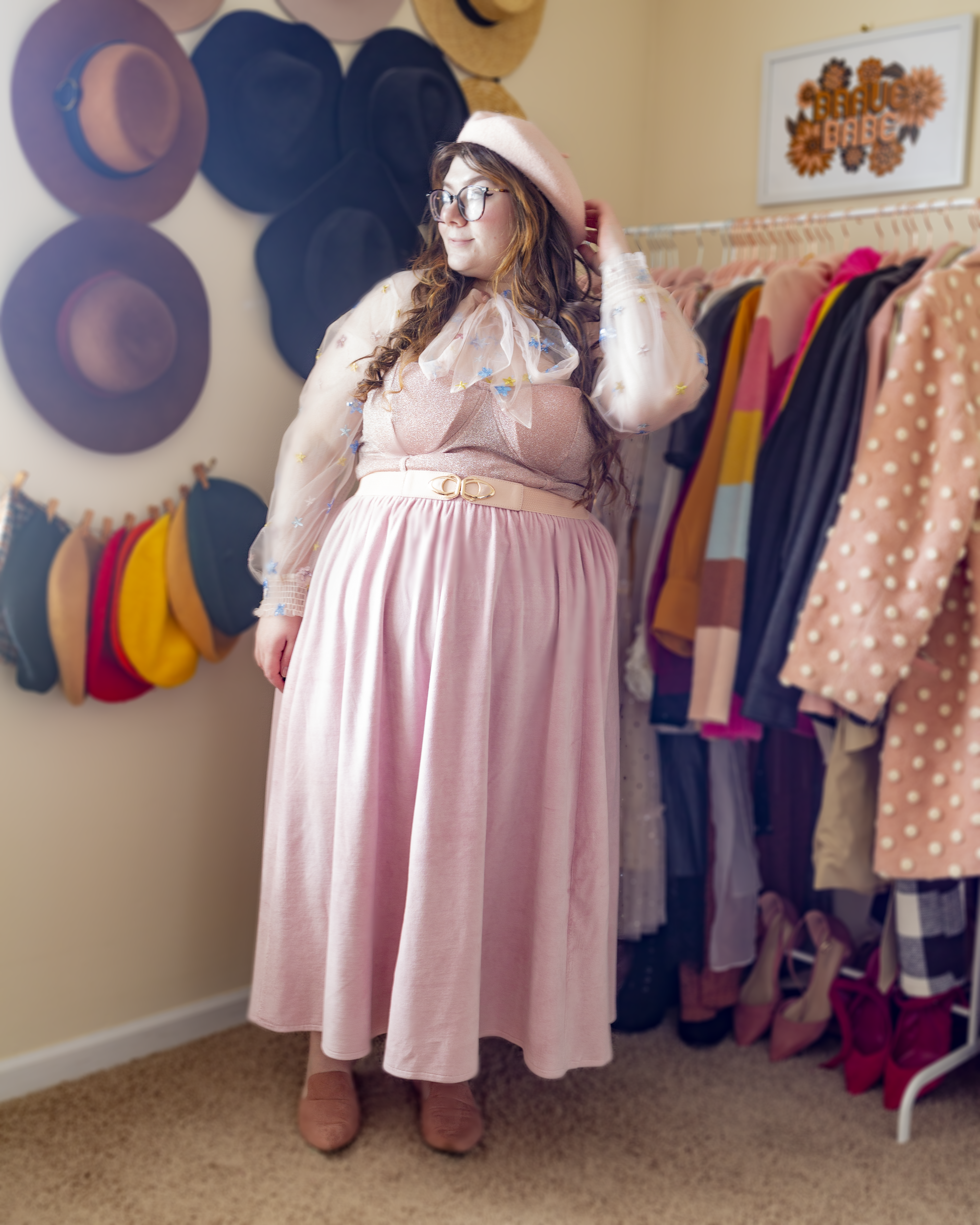 An outfit consisting of a beige wide brim hat, sheer white sheer bow blouse with pastel pink, yellow and blue sequin flowers, tucked into pastel pink velour midi skater skirt, and pink mules.