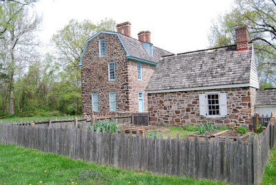 Small stone kitchen building with a garden surrounded by rough wooden fencing is in the foreground, the two-story stone Keith House with blue trim around doors and windows stands behind