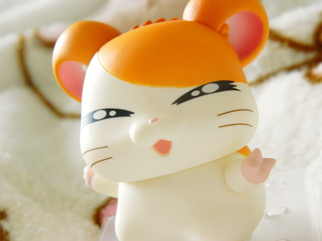 A photo showing a figure of an orange and white anime hamster character called Hamtaro. He is smiling with closed eyes and has his right hand held up in the peace sign.