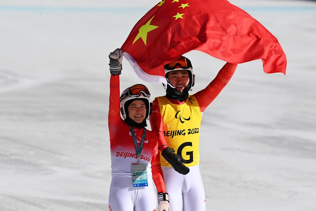 Atleta chinesa, junto de seu atleta guia, carregam a bandeira do país durante a comemoração. Ambos vestem uma malha branca e vermelho, sengo que o guia veste um colete amarelo com a letra G por cima