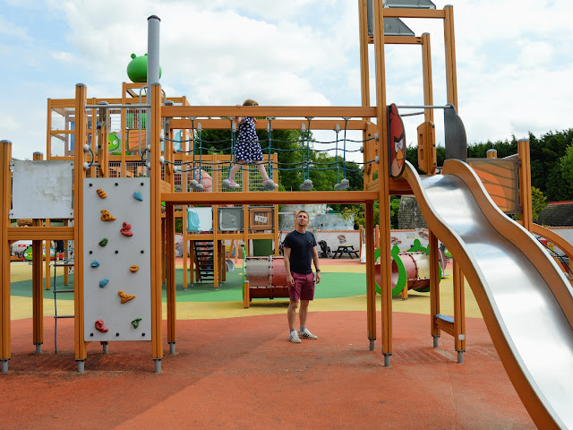 Image of the play equipment in the Angry Birds area of Sundown Adventureland. The image shows a young girl walking across a rope bridge within a wooden framed climbing frame. To the left of the rope bridge is a climbing wall and to the right is a wavy metal slide. The floor is rainbow coloured protective rubber flooring.
