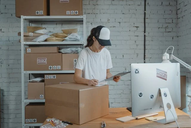 Woman writing down on a Carton Box while looking at a Clipboard In Her Reselling Business