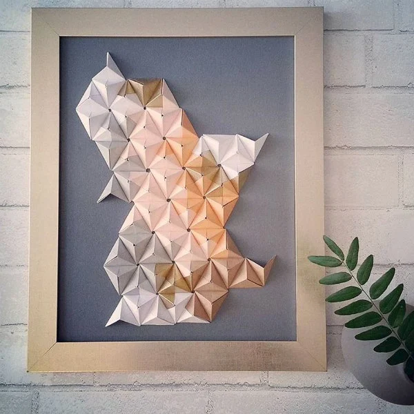 3D origami art in light, neutral shades with light wood frame displayed on wall