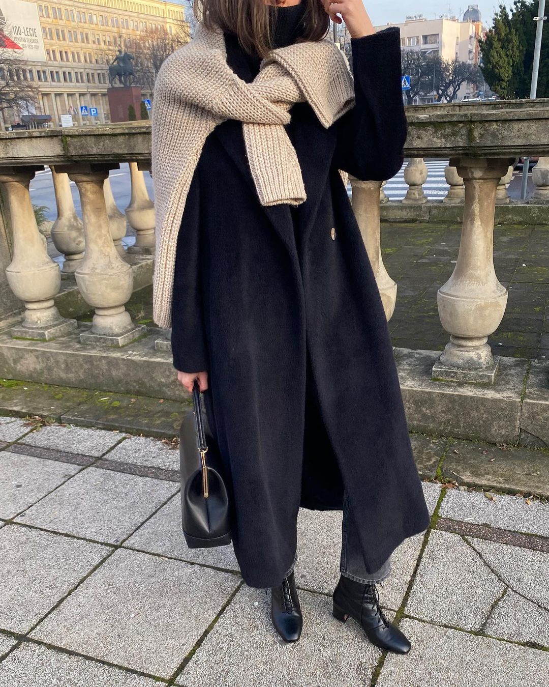 Chic Winter Outfit Idea — Instagram Style From @jestem_kasia Kasia Szymków in with Long Black Coat, Beige Sweater Tied Over The Shoulders, Faded Black Jeans, and Square Toe Boots