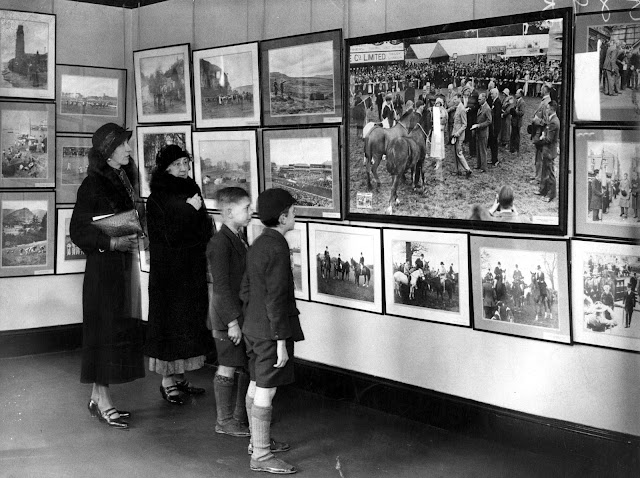 Two women and two young boys, all very smartly dressed, viewing a photography exhibition. Lots of framed photographs are hung together very closely, completely covering the walls.