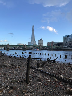 A picture showing the view over the pebble strewn foreshore of the River Thames to the Shard building in London.  There are various posts of rotting wood sticking up out of the mud and pebbles.  Photograph by Kevin Nosferatu for the Skulferatu Project