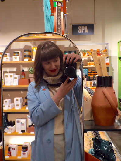 Ellie taking a selfie in the new gold edged mirror beside the makeup section, showing the top half of her outfit, being a cream roll-neck chunky knit jumper, dusky blue pencil skirt and blue long mac. Red lips and hair in curls, with camera to hand and a little booby lady pot sits on the makeup shelf for display.