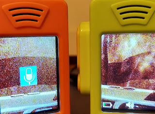 The orange walkie talkie screen is showing a blue and white mic icon in the middle of the screen. The Green walkie-talkie's screen is showing the battery icon in the bottom left corner. The battery is showing one green bar. The speaker icon is a white icon in the  bottom center.