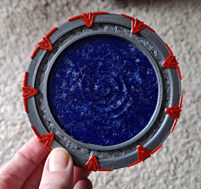 A plastic coaster that looks like a stargate, 3d-printed in PETG with silver gate ring, red accents, and translucent blue event horizon.