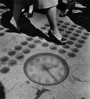 black and white photograph of feet of a woman and man walking by a clock embedded in the sidewalk in NYC, 1947