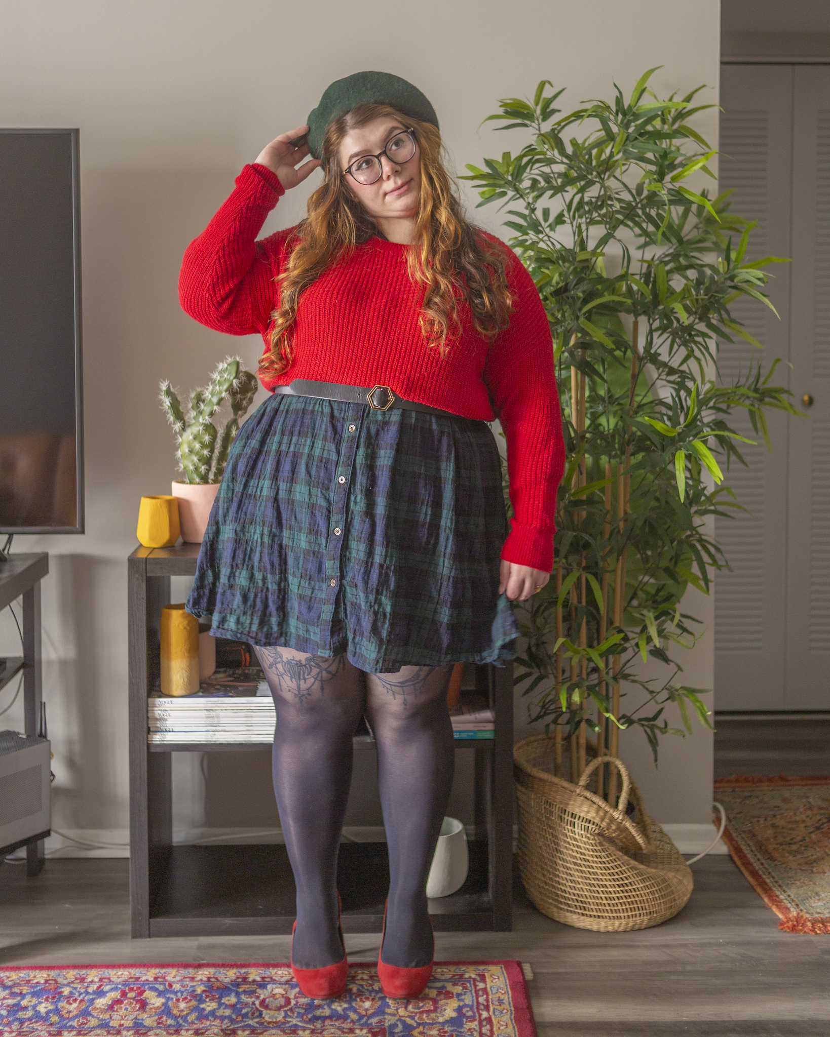 An outfit consisting of a red cropped sweater over a green plaid shirt smock dress, black tights and black Chelsea boots.