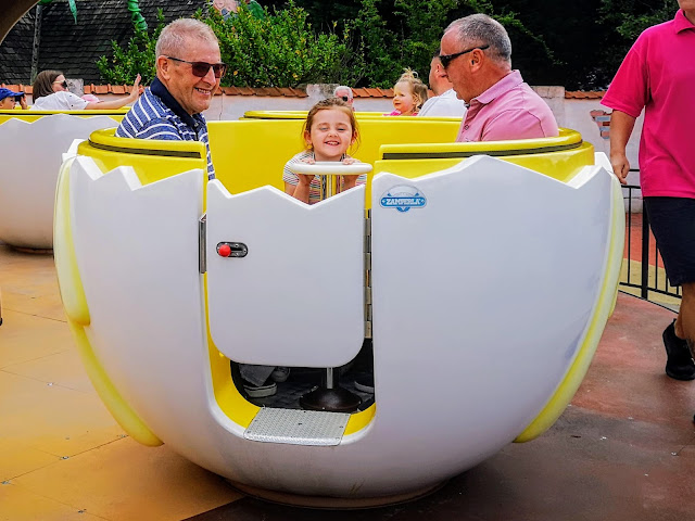 Image of the egg yolk ride at Sundown Adventureland. The image shows a young girl and both her grandads sat inside a ride that looks like half a cracked egg shell. The girl is smiling and has her hands on the central pedestal that is used to make the egg shell spin.