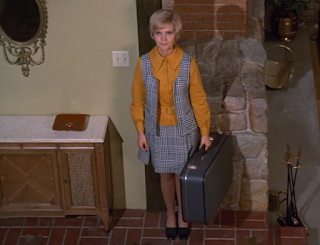 Carol Brady waits at the bottom of the stairs holding a suitcase.