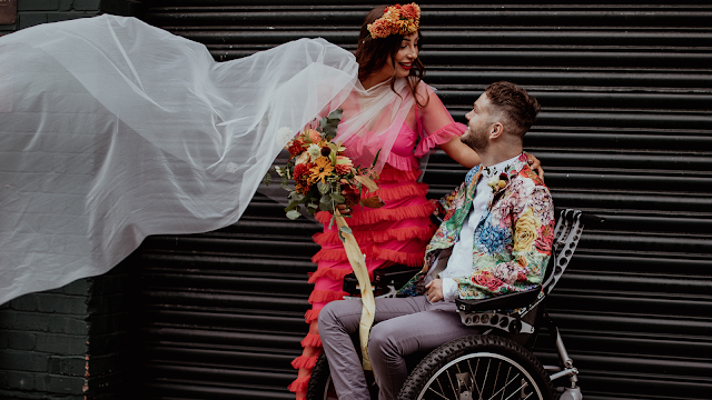 a man in a wheelchair wearing a patterned jacket looks lovingly at a woman in a bright pink dress and a flowing veil.