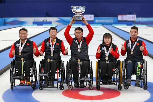 Cinco atletas chineses posam para fotos em suas cadeiras de rodas em pista de curling. O atleta do meio levanta a taça da Copa do Mundo de curling