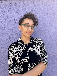 a mixed-raced Black & white person stands in front of a lilac wall. They have short curly hair and translucent green glasses. They are wearing a black button-down shirt with a white floral design. They’re looking off to the side, smirking.