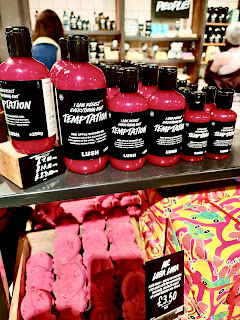 A photo of a row of cylindrical clear plastic bottles filled with red liquid with black labels that say I can resist anything but temptation in shower gel in white font with black cylindrical bottle lids with a black rectangular portrait card with temptation shower gel in white font on a light brown rectangular shelf  on a bright background