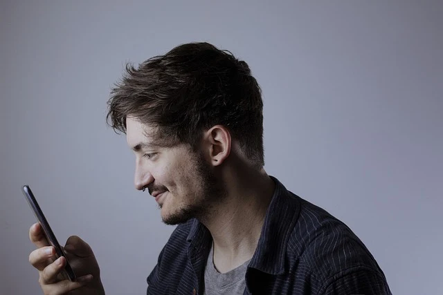 A man with short, slightly messy hair and a trimmed beard smiling while looking at his smartphone. He is wearing a dark shirt, and the background is minimalistic with a soft gray gradient.