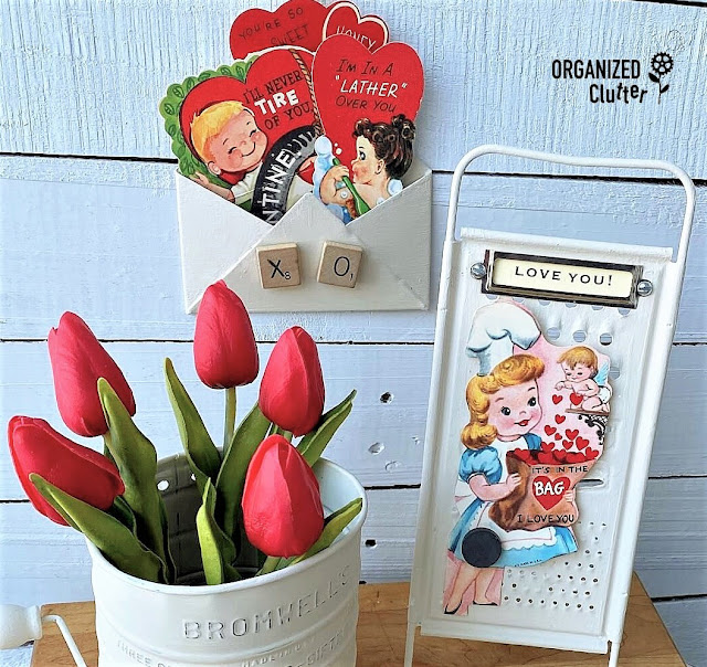 Photo of a kitchen grater with easel back, a Bromwell sifter, and a Dollar Tree mini metal envelope painted in Drop Cloth Chalk Paint. Items used in a Valentine's Day vignette with vintage Valentines and faux flowers.