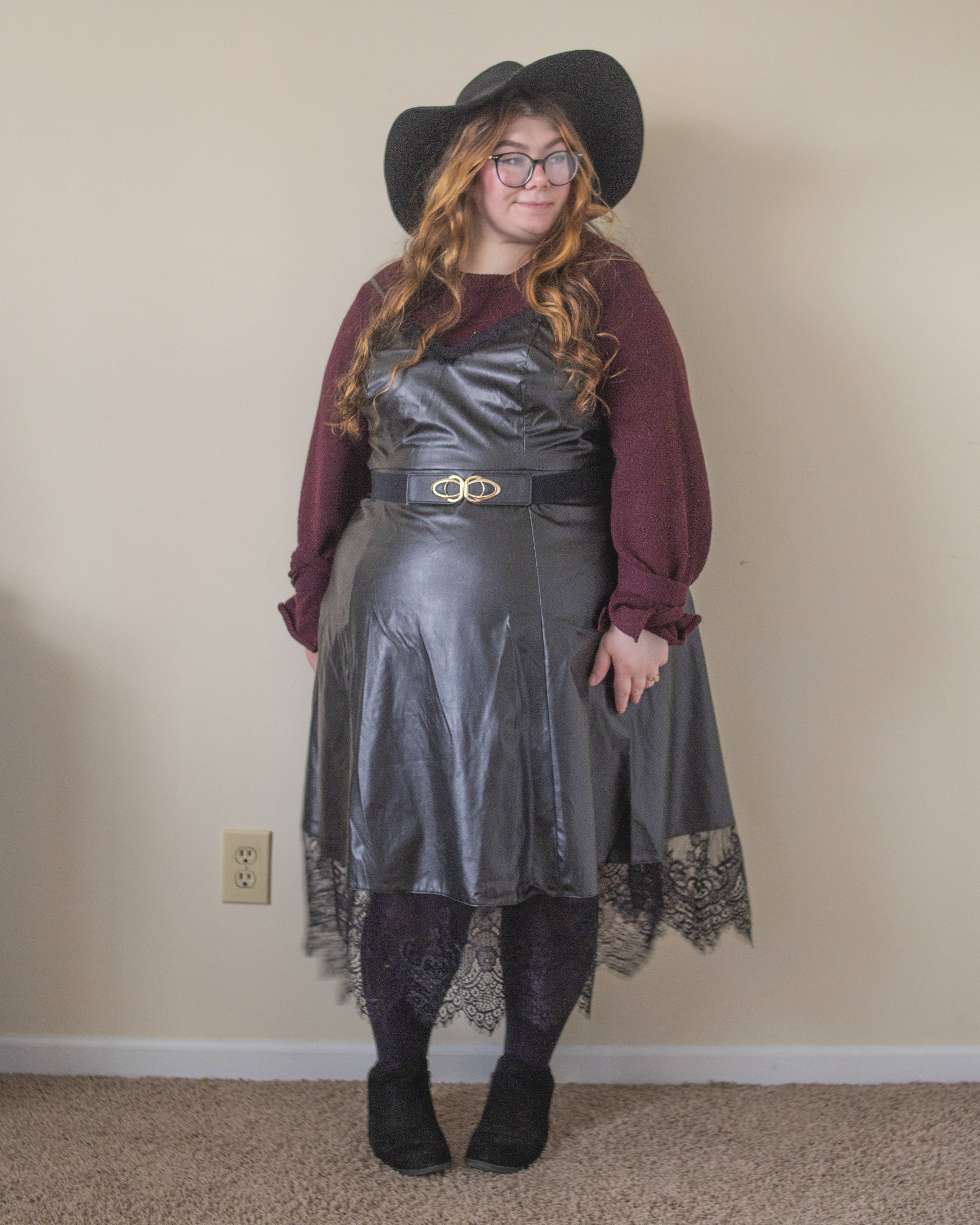 An outfit consisting of a berry knit sweater with ties around the wrists under a black leather midi dress with black lace trim, black tights and black boots.