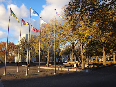 Autumn 2015 photo of the Garden of the Provinces and Territories park, with six provincial flags on poles, and a zen-like water feature in the treed area of the park. The Library and Archives Canada building is in the background.