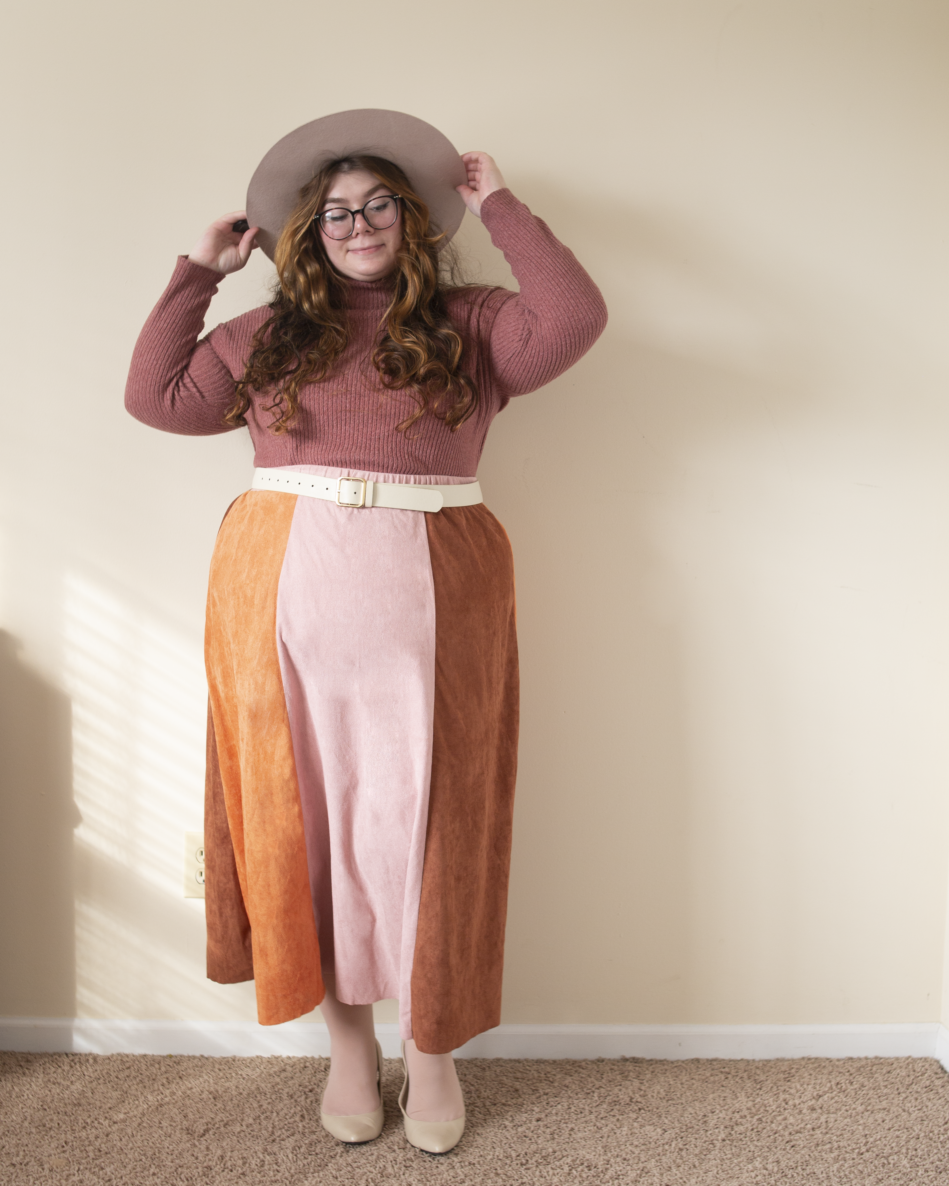 An outfit consisting of a muted pink turtleneck tucked into a burnt orange, cognac brown and pastel pink panel suede midi skirt and brown midi boots.