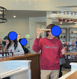 A man is holding aloft a flyte of maple syrups. He's standing in the middle of a wooden counter, that circles around him. On the other side of the counter are people with their own flutes, listening to him. All their faces have been obscured.