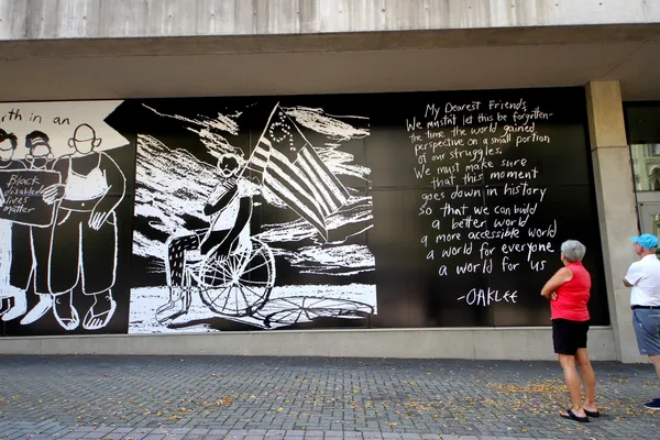 Three pieces of large scale art on the exterior of a building. Left side partial shows three people wearing masks, holding a sign that says "Black disabled lives matter". Middle piece shows person in wheelchair, holding a flag that is similar to the American flag except that the stars have been moved into the shape of the "wheelchair accessibility" symbol. Right panel has text: "My Dearest Friends, We mustn't let this be forgotten - the time the world gained perspective on a small portion of our struggles. We must make sure that this moment goes down in history so that we can build a better world a more accessible world a world for everyone a world for us  -- Oaklee". Two passersby are in lower right corner of image, looking up at the exhibit.