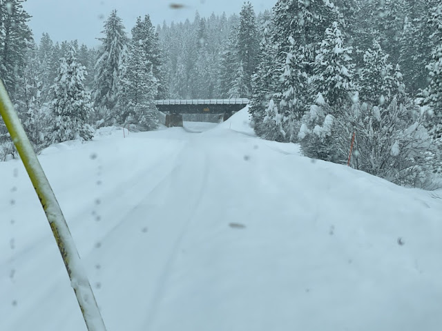 A mountain road completely covered with snow; an overpass crossing the road is also white with snow.