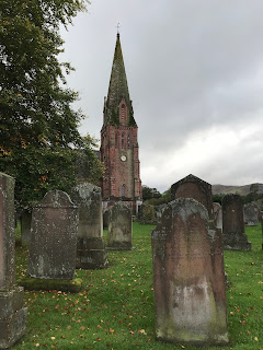 A photograph of several old and worn gravestones in Penpont Churchyard, with the Parish Church in the background.