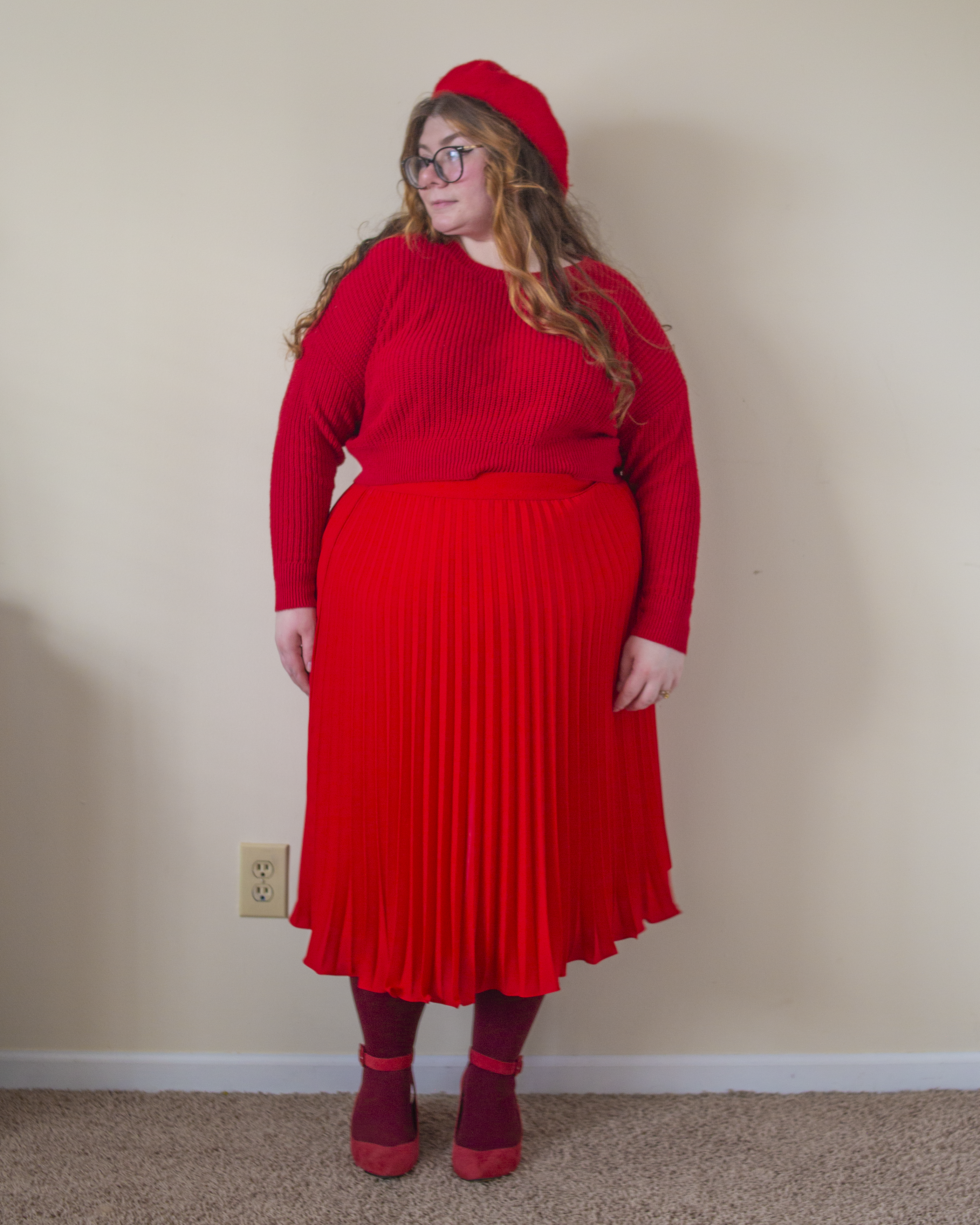 An outfit consisting of a red beret, red sweater half tucked into a red pleated midi skirt with maroon tights with red ankle strap heels.