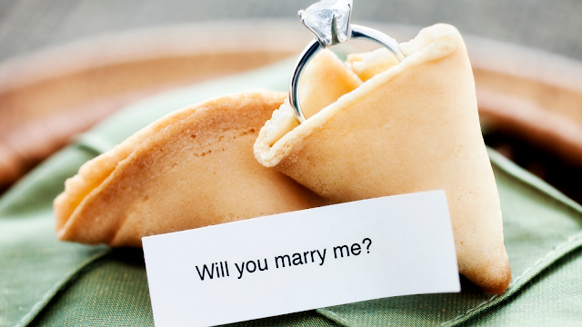 An engagement ring is nestled inside a fortune cookie and the note reads "will you marry me?".