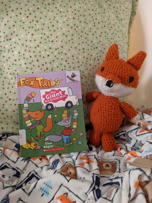 ID: a photo by Erin Liu shows the book Fox Tails: a Giant Ice Cream Mess and a crocheted fox, made by Erin Liu, on top of a woodland themed sheet with foxes on it..