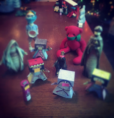 ID: Magi from various nativities, a red bear, and a Granddaddy napkin holder face the camera, led by a magi in a purple dress and no facial hair. All have masks. In the background is the shelter where the holy family is living with a penguin and small red bear as well as the inhabitants.