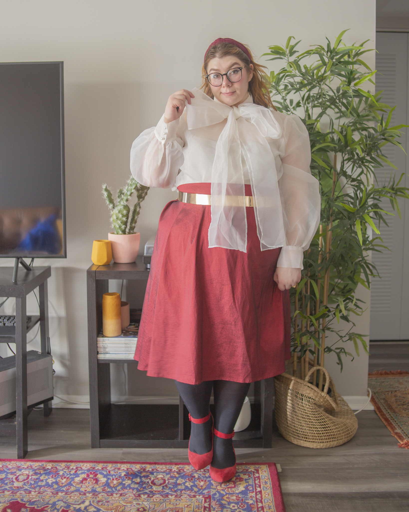 An outfit consisting of a sheer white pussy bow blouse under a white sweater, tucked into a red metallic midi skirt and red ankle strap heels.
