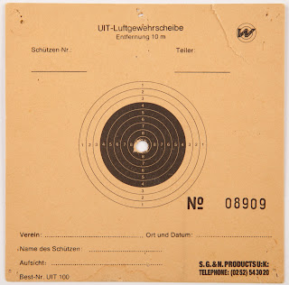 A square cardboard target shot with all 10 shots through the central ring