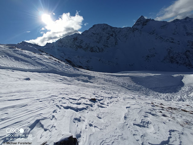 Im Vordergrund eine massiv vom Wind geprägte Schneeoberfläche, im Hintergrund ein Lawinenanriss samt Ablagerung. Person konnte unverletzt entkommen. Leppleskogel - Osttirol (Foto: 14.12.2021)